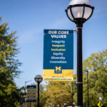 "University of Michigan street banner displaying core values: Integrity, Respect, Inclusion, Equity, Diversity, and Innovation, with information on how to learn more at culturejourney.umich.edu. The banner is hanging from a lamppost on a sunny day, surrounded by trees.