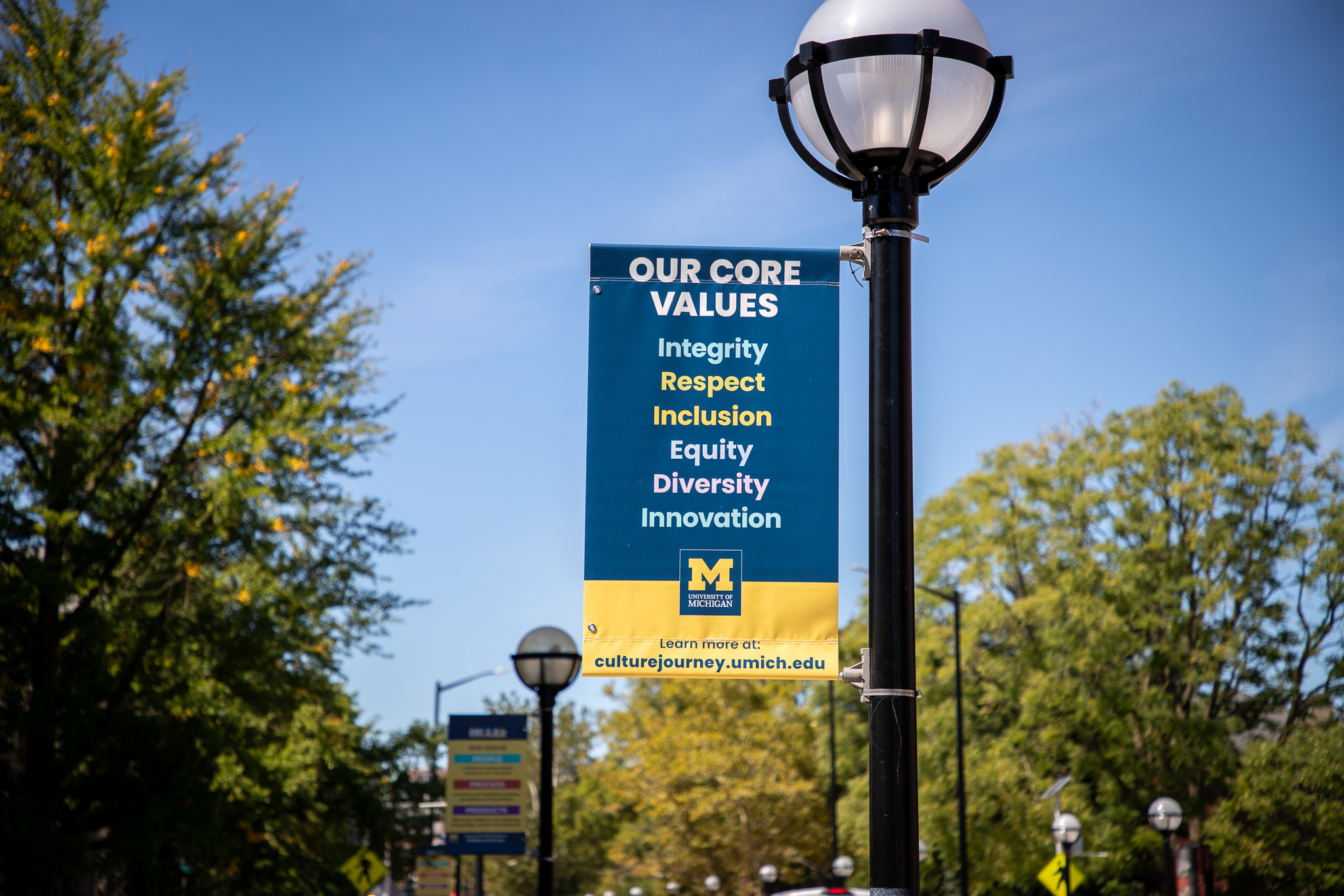 "University of Michigan street banner displaying core values: Integrity, Respect, Inclusion, Equity, Diversity, and Innovation, with information on how to learn more at culturejourney.umich.edu. The banner is hanging from a lamppost on a sunny day, surrounded by trees.