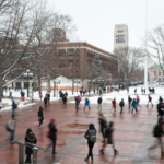 A winter scene on the University of Michigan campus, featuring the bustling Diag with students walking on brick pathways surrounded by snow-covered trees and buildings, including the iconic Burton Memorial Tower in the background.