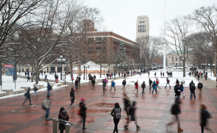 A winter scene on the University of Michigan campus, featuring the bustling Diag with students walking on brick pathways surrounded by snow-covered trees and buildings, including the iconic Burton Memorial Tower in the background.