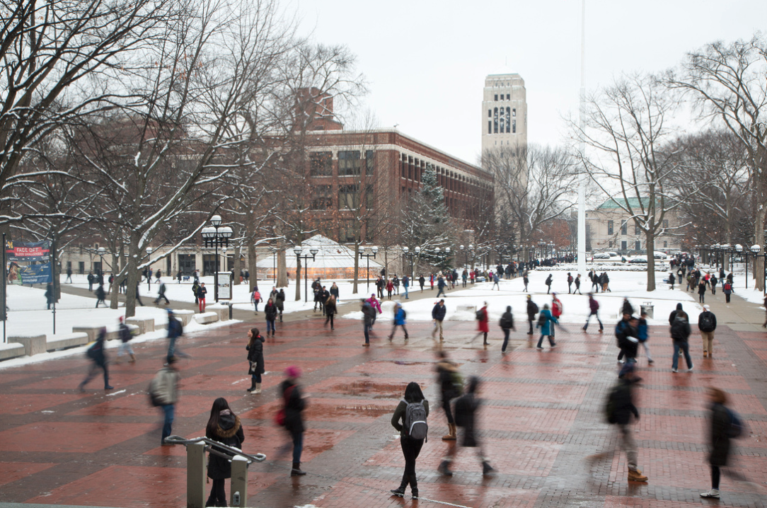 A winter scene on the University of Michigan campus, featuring the bustling Diag with students walking on brick pathways surrounded by snow-covered trees and buildings, including the iconic Burton Memorial Tower in the background.